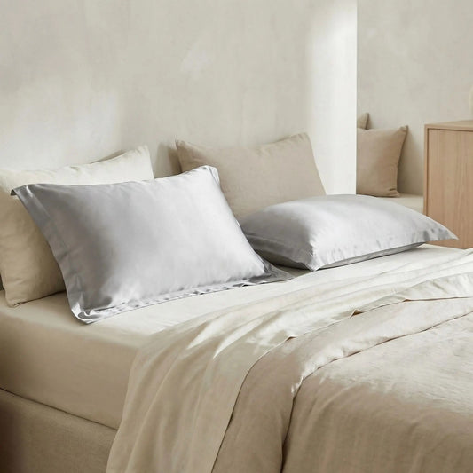 Taie d’oreiller en soie de mûrier 30 mommes gris argenté posée sur un lit en bois clair avec linge de lit clair rendu luxueux et lumineux