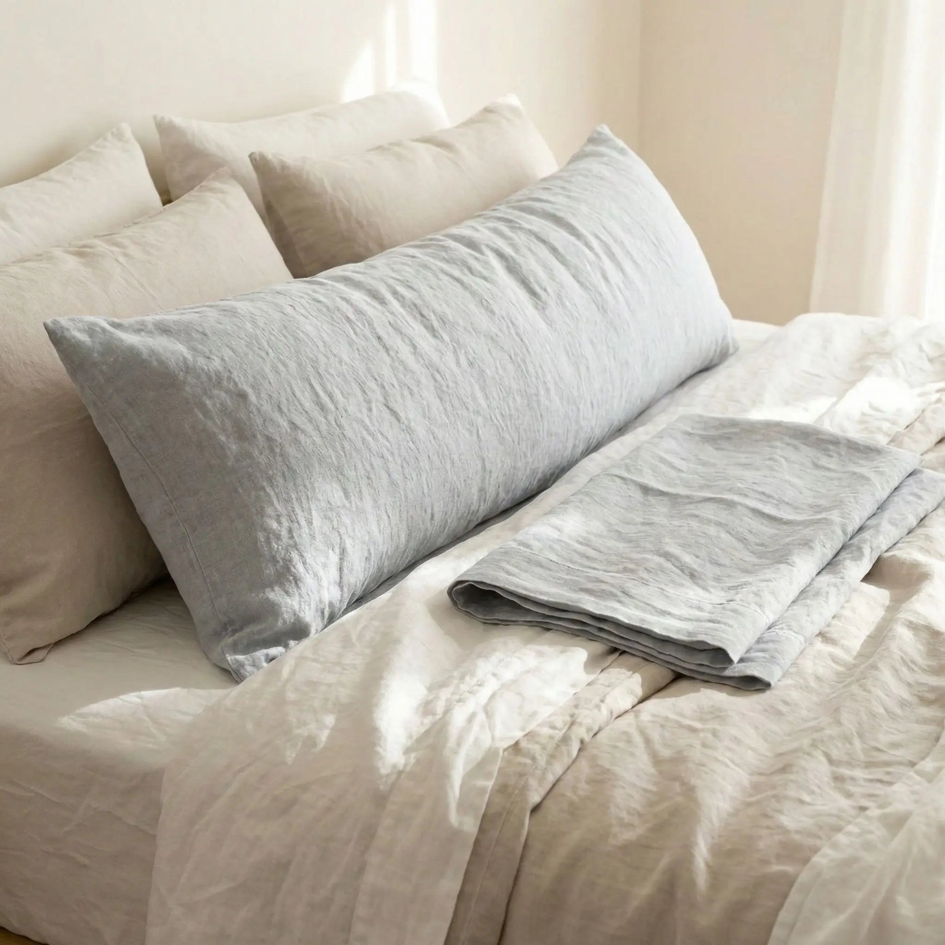 Taie de traversin en lin gris clair posée sur un lit en bois clair avec linge de lit blanc cassé ambiance lumineuse et naturelle
