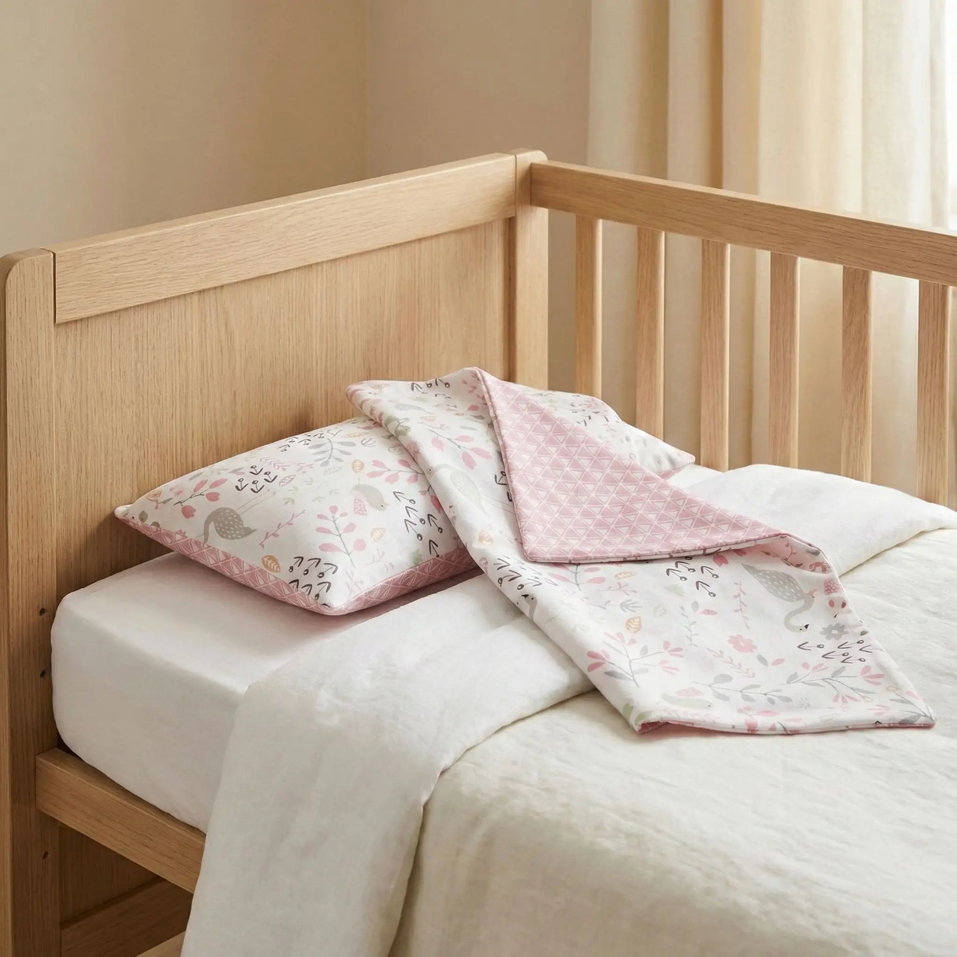 Taies d’oreiller bébé en coton à motif fleurs roses posées sur un lit bébé en bois clair avec linge de lit blanc cassé