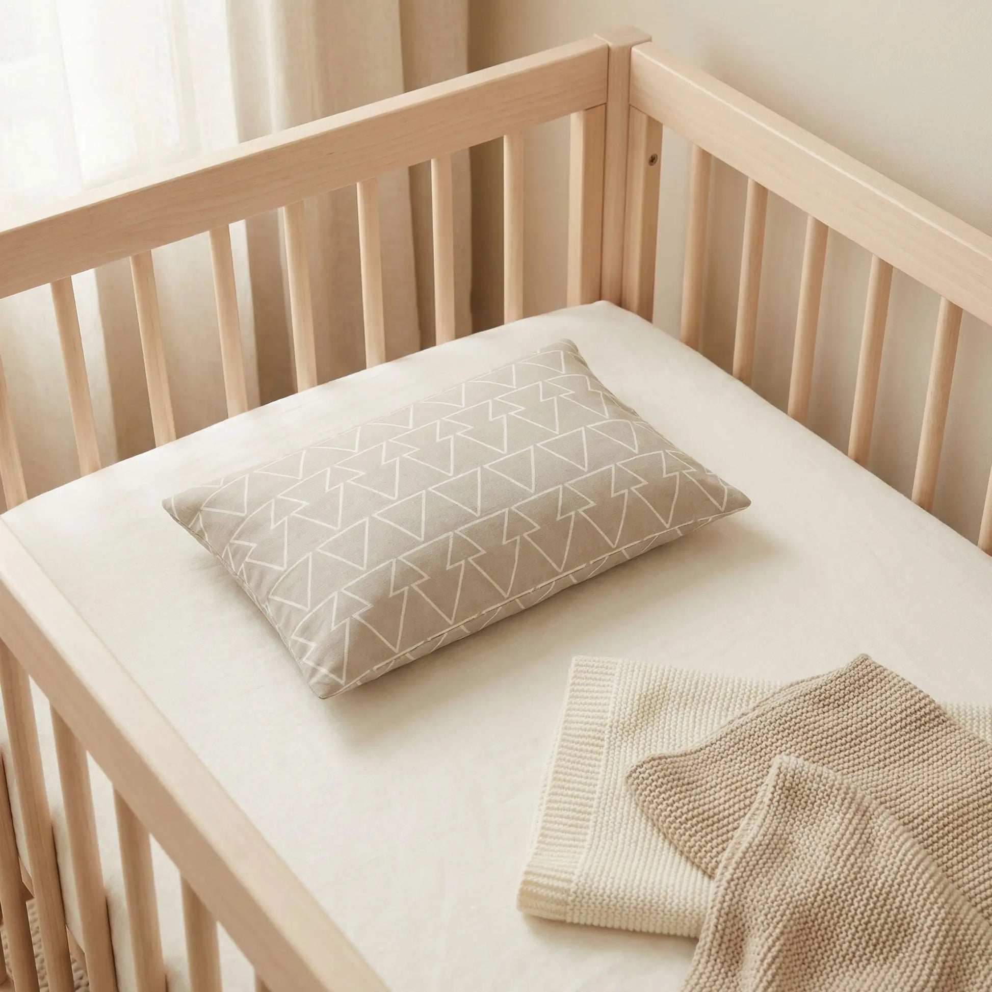 Taies d’oreiller bébé en coton à motif triangles posées sur un lit bébé en bois clair avec linge de lit blanc cassé