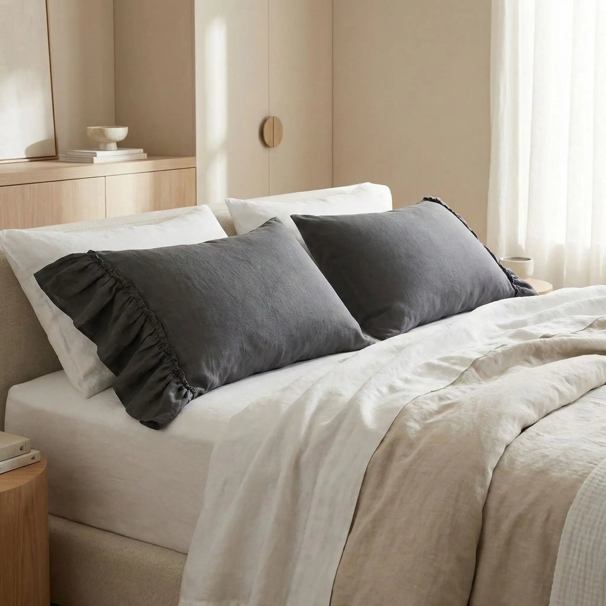 Taies d’oreiller en lin lavé gris foncé posées sur un lit en bois clair avec linge de lit blanc cassé