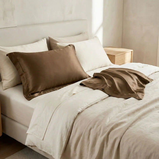 Taies d’oreiller en satin marron posées sur un lit en bois clair avec linge de lit blanc cassé ambiance lumineuse et naturelle