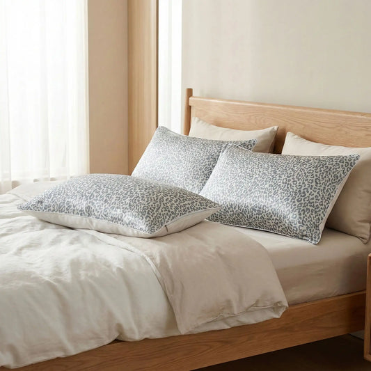 Taies d’oreiller en soie grises posées sur un lit en bois clair avec linge de lit blanc cassé