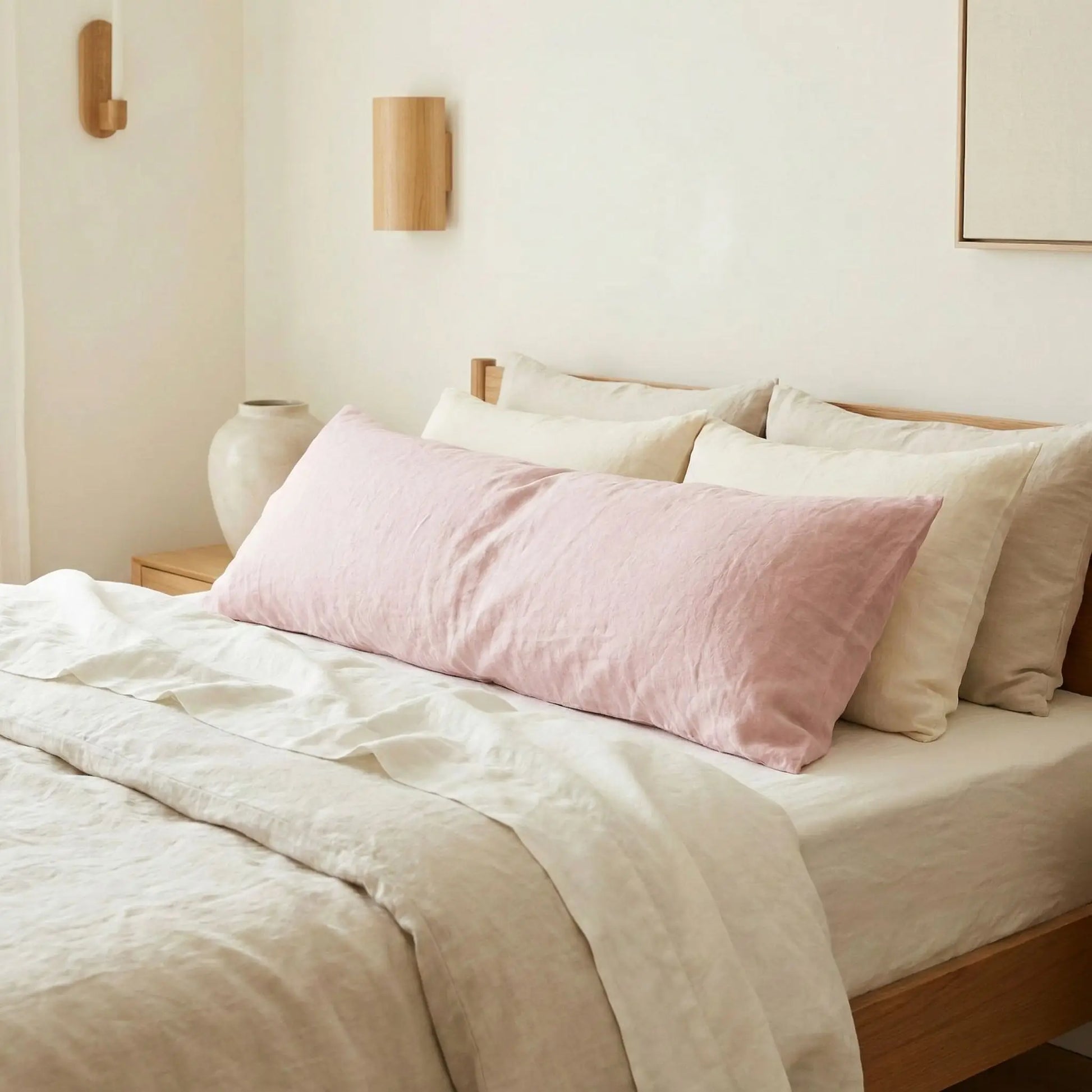 Taies de traversin en lin rose posées sur un lit en bois clair avec linge de lit blanc cassé