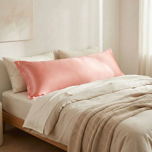 Taies de traversin en satin rose pêche satinée posées sur un lit en bois clair avec linge de lit blanc cassé